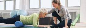 Pokaż zdjęcie: Starsza pani wykonuje ćwiczenia rehabilitacyjne. Leży na brzuchu na wałku gimnastycznym. Obok niej klęczy młoda rehabilitantka. 