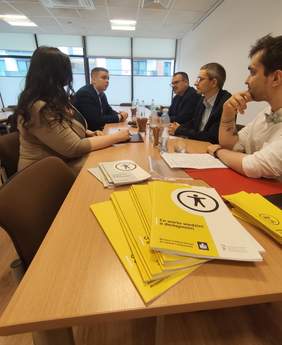 Pokaż zdjęcie: Grupa osób siedzi przy stole podczas spotkania, na pierwszym planie widoczne są broszury informacyjne dotyczące dostępności.