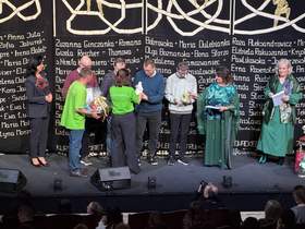 Scena teatru. Na tle ciemnej kurtyny stoi kilka osób - kobiety i mężczyźni. Cześć osób wręcza, część odbiera nagrody.