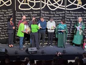 Pokaż zdjęcie: Scena teatru. Na tle ciemnej kurtyny stoi kilka osób - kobiety i mężczyźni. Cześć osób wręcza, część odbiera nagrody.