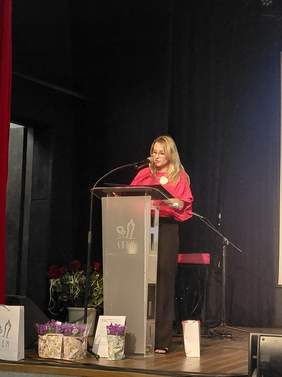 Pokaż zdjęcie: Kobieta na scenie przemawia do mikrofonu przy jasnoszarej mównicy. Scena jest oświetlona od przodu, a w ciemnym tle za prelegentką znajduje się kurtyna. Obok mównicy, na podłodze, stoją ozdobne torebki prezentowe oraz kwiaty w doniczkach.