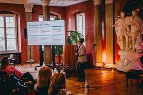 Pokaż zdjęcie: Sala konferencyjna z publicznością siedzącą na krzesłach i zwróconą w stronę sceny. Na środku stoi osoba przemawiająca do publiczności i trzyma mikrofon. Po lewej stronie sceny ustawiony jest ekran projekcyjny i wyświetlony jest na nim slajd. Ściany mają ciepły, czerwony kolor, a oświetlenie w odcieniach pomarańczowo niebieskich podkreśla kolumny. Na ekranie projekcyjnym widnieje tekst: „Kto potrzebuje AAC w szkole”.