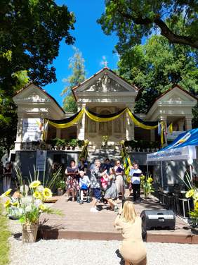 Pokaż zdjęcie: Na zewnątrz, na scenie trwa występ wokalny, grupa 8 osób tworzy chór, na scenie tańczy dziewczyna z chłopakiem.