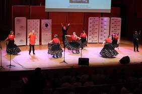 Pokaż zdjęcie: Na scenie Sali Koncertowej trwa występ osób z niepełnosprawnościami. Artyści mają na sobie cygańskie stroje. Tańczą i wykonują różne obroty.  