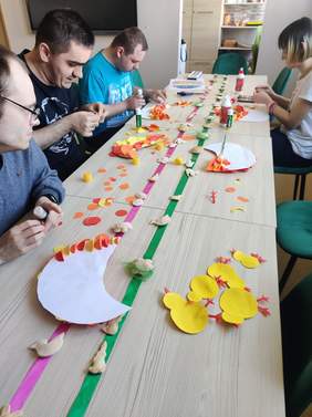 Pokaż zdjęcie: kilka osób siedzi przy prostokątnym stole na którym są umieszczone prace plastyczne z gliny oraz wycinanki o charakerze wielkanocnym