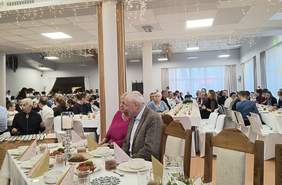 Pokaż zdjęcie: Zdjęcie przedstawia dużą salę, w której odbywa się wydarzenie, prawdopodobnie uroczysty obiad lub kolacja. Uczestnicy siedzą przy elegancko nakrytych stołach, przyozdobionych świecami i dekoracjami. W tle widać lampki i ozdoby na suficie, które dodają świątecznej atmosfery.