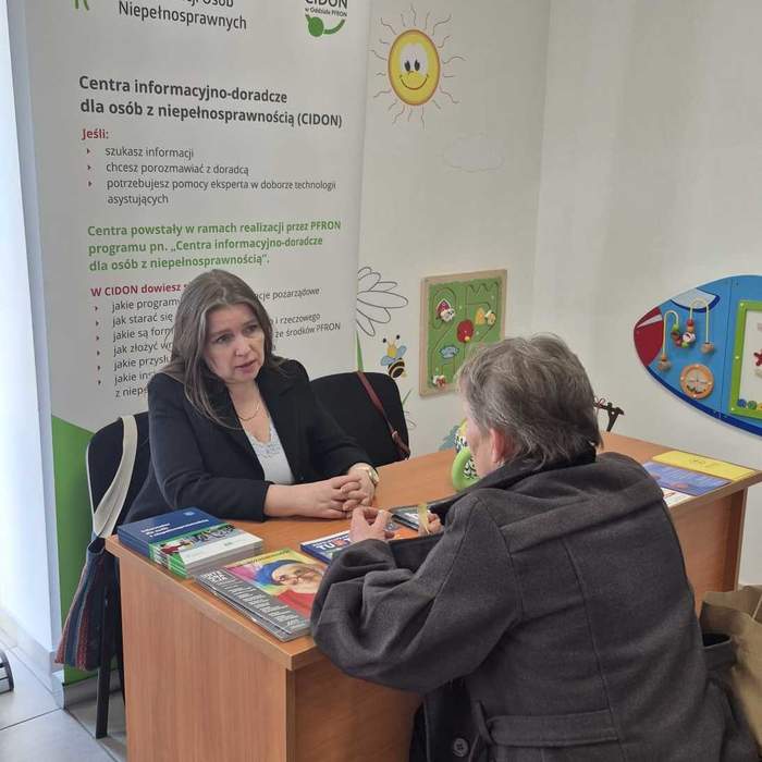 Pokaż zdjęcie: Spotkanie przy biurku w punkcie informacyjno-doradczym PFRON, gdzie dwie osoby rozmawiają przy materiałach informacyjnych, na tle roll-upu i kolorowych dekoracji ściennych.
