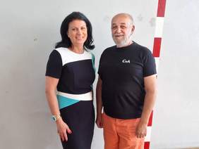 Pokaż zdjęcie: Uśmiechnięci kobieta i mężczyzna stoją obok siebie na tle jasnej ściany. Kobieta ma ciemne, rozpuszczone włosy i nosi elegancką sukienkę w kolorach czerni, bieli i turkusu, a mężczyzna ma krótko przystrzyżoną siwą brodę i ubrany jest w czarną koszulkę z napisem „CeA” oraz pomarańczowe spodnie.