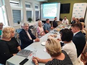 Pokaż zdjęcie: Przy prostokątnym stole siedzą kobiety i mężczyźni. Rozmawiają ze sobą. W tle widać tablicę multimedialną z wyświetloną prezentacją. 