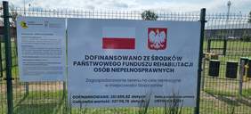 Pokaż zdjęcie: Na ogrodzeniu zawieszona jest duża tablica informacyjna. Zawarte są na niej informacje dotyczące budowy placu zabaw dla dzieci. 