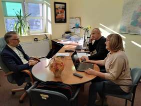 Pokaż zdjęcie: Przy stole siedzi kobieta i dwóch mężczyzn. Rozmawiają ze sobą i patrzą na siebie. Przed panią leży otwarty laptop. 