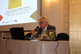 Pokaż zdjęcie: Przy stole konferencyjnym siedzi mężczyzna w garniturze i okularach, przemawia do mikrofonu. W tle zdjęcie prezentacji komputerowej