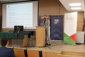 Pokaż zdjęcie: Przy mównicy mężczyzna z brodą w jasnym garniturze, zdjęcie z auli