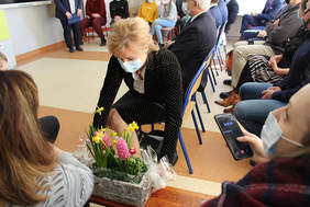 Pokaż zdjęcie: Kobieta siedząca na krześle, pochyla się i odstawia na podłogę kwiaty, w tle inne osoby siedzące na krzesłach