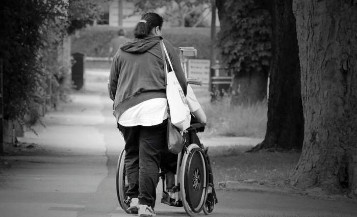 Pokaż zdjęcie: Opiekun prowadzi osobę niepełnosprawną na wózku inwalidzkim