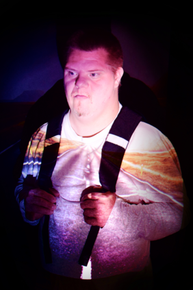 Pokaż zdjęcie: Na czarnym tle widać zdjęcie postać mężczyzny w ujęciu do połowy ciała, mężczyzna jest tęższy, ma widoczny brzuszek, ubrany jest w białą podkoszulkę i spodnie z grubymi czarnymi szelkami za które trzyma się rękami. Na tle koszulki odbija się droga z widocznym poboczem i malatura pasów drogowych idących pionowo przez środek.