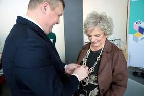 Pokaż zdjęcie: Mężczyzna w garniturze przypina medal do klapy marynarki starszej kobiety w brązowej marynarce