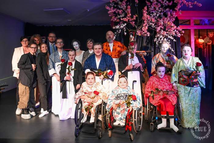 Pokaż zdjęcie: Grupowe zdjęcie aktorów na scenie, ustawione pod rozłożystym, sztucznym drzewem kwitnącej wiśni (sakura). Scena oświetlona jest na niebiesko-fioletowo. Aktorzy stoją i siedzą w dwóch rzędach, ubrani w stroje inspirowane kulturą japońską: kimona, niektóre osoby mają białą charakteryzację twarzy. Trzy aktorki w pierwszym rzędzie siedzą na wózkach inwalidzkich, a każda z osób na pierwszym i środkowym planie trzyma w dłoni jedną czerwoną różę.