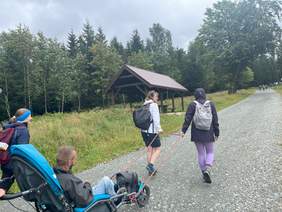 Pokaż zdjęcie: Dwie osoby ciągną za sznur terenowy wózek inwalidzkim, na którym siedzi osoba z niepełnosprawnością. Obok wózka idzie kobieta.