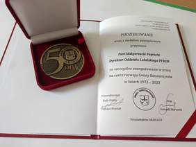 Pokaż zdjęcie: W czerwonym pudełku znajduje sie złoty medal z treścią 50 lat gminy Konstantynów, obok leżą podziękowania dla Pani Małgorzaty Paproty za szczególne zaangażowanie i pracę na rzecz rozwoju gminy Konstantynów.