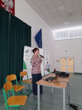 Pokaż zdjęcie: Prelegentka stojąca przy stole, w ręku trzyma mikrofon, patrzy w kierunku słuchaczy.