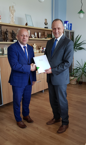 Dwóch mężczyzn w garniturach, stoją, ściskają sobie dłonie, przekazują dobie dokumenty. Uśmiechają się.