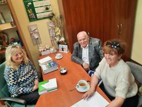 Pokaż zdjęcie: Przy podłóżnym stole siedzą trzy osoby: dwie kobiety i jeden mężczyzna. Wszyscy się uśmiechają. Na stole leżą różne dokumenty, informatory, poradniki i filiżanki z herbatą. 