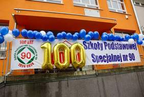Pokaż zdjęcie: Na budynku szkoły zawieszona jest dekoracja z okazji 100-lecia placówki. Wiszą transparenty i złote balony. 