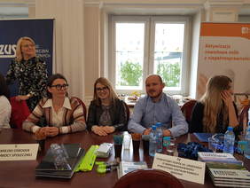 jedna kobieta stoi, trzy kobiety i jeden mężczyzna siedzą na tle okna i baneru, uśmiechają się