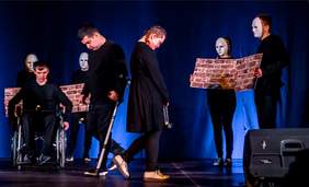 Pokaż zdjęcie: scena w kolorze granatowym, na scenie stoją osoby ubrane na czarno jedna z nich jest na wózku inwalidzkim, cztery z nich są w maskach i trzymają mur z cegieł