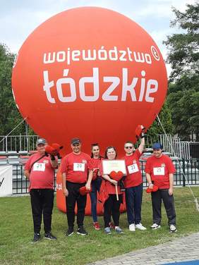 Pokaż zdjęcie: W rzędzie stoi sześć osób: dwie kobiety i czterech mężczyzn. Wszyscy mają czerwone koszulki i trzymają czerwone pluszowe serduszka. Nad nimi jest wielki czerwony balon z napisem "Województwo łódzkie". 