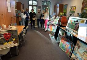 Pokaż zdjęcie: W pokoju biurowym stoi kilka osób. Dokoła nich rozłożone są prace plastyczne.