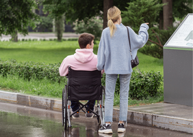 Pokaż zdjęcie: Tyłem stoją dwie osoby: chłopak siedzący na ręcznym wózku inwalidzkim i stojąca obok niego dziewczyna. W tle widać zieleń: drzewa, krzewy i trawę. 