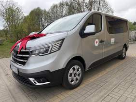 Pokaż zdjęcie: Srebrny bus do przewozu osób z niepełnosprawnościami stoi na parkingu. Na masce widoczna duża czerwona kokarda, a na drzwiach oznaczenia organizacji i Logo PFRON.