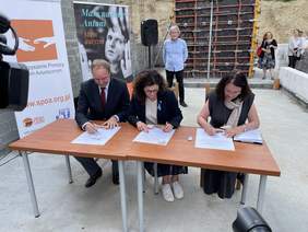 Pokaż zdjęcie: Mężczyzna i dwie kobiety siedzą przy jednym stole. Trzymają w rękach długopisy i podpisują dokumenty. Wszyscy galowo ubrani.