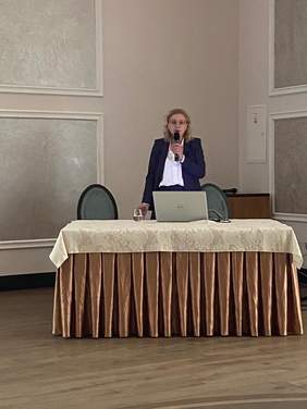 Pokaż zdjęcie: Kobieta blondynka w ciemnej marynarce stoi przy stole z laptopem, wygłasza prelekcję.