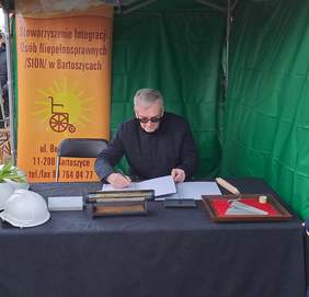 Pokaż zdjęcie: Mężczyzna podpisuje akt erekcyjny przy stole z ciemnym obrusem. Za nim kolorowy baner z napisem ,,Stowarzyszenie Integracji Osób Niepełnosprawnych SION w Bartoszycach’’