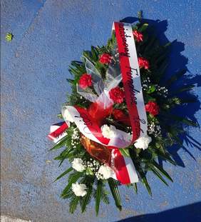 Pokaż zdjęcie: Na asfalcie leży wiązanka kwiatów. Składa się ona z biało-czerwonych goździków. Przepasana jest szarfą z napisem: Państwowy Fundusz Rehabilitacji Osób Niepełnosprawnych. 