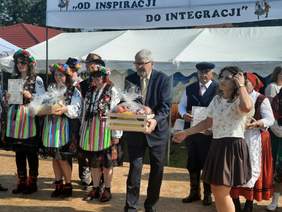 Pokaż zdjęcie: Dyrektor Łódzkiego Oddziału PFRON trzyma w dłoniach upominkowy prezent. Wokół niego są osoby ubrane w ludowe stroje. Nad głowami ludzi wisi transparent z hasłem: "Od inspiracji do integracji". 