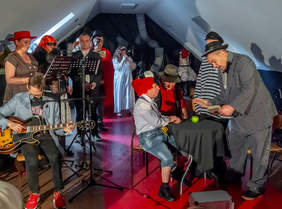 Pokaż zdjęcie: Na scenie trwa występ artystyczny osób z niepełnosprawnościami. Aktorzy mają na sobie przeróżne przebrania. Po lewej stronie gra zespół muzyczny. 