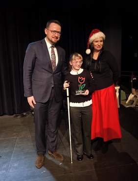 Pokaż zdjęcie: Troje osób pozuje do zdjęcia na tle ciemnej kurtyny. Po lewej stronie stoi mężczyzna w okularach i garniturze. W środku znajduje się młoda osoba trzymająca białą laskę oraz małą statuetkę. Po prawej stoi kobieta w czarnej bluzce, długiej czerwonej spódnicy i czapce świętego Mikołaja, z ręką opartą na ramieniu laureatki.