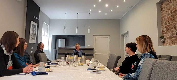 W jasnej, nowoczesnej sali, przy stole siedzi kilka osób, a na szczycie kobieta, ubrana w mundur służby więziennej. Na stole stoją termosy, szklanki i talerzyki z ciastem.