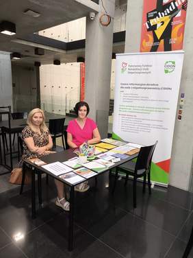 Pokaż zdjęcie: Dwie kobiety siedzą przy stoliku na którym leżą ulotki, w tle baner z logo funduszu i CIDON.