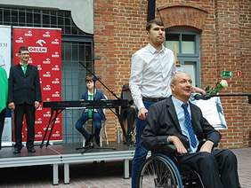 Mężczyzna na wózku inwalidzkim mówi do mikrofonu, za nim asysten w bialej koszuli, za nimi małą scena a na niej dwie osoby, wszystko na tle ściany z cegły