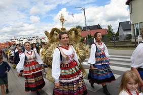 Pokaż zdjęcie: Przemarsz dużej grupy osób, kobiety w ludowych strojach trzymają wieniec dożynkowy 