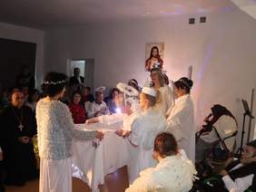 Pokaż zdjęcie: Zdjęcie podczas występu, artyści przebrani za anioły trzymają w ręku biały obrus. W tle widać gości siedzących na krzesełkach oraz biskupa.