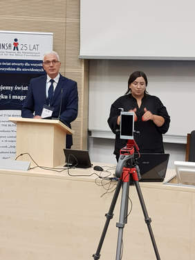 Pokaż zdjęcie: od lewej mężczyzna w garniturze stoi przed mikrofonem za mównicą, obo kobieta ubrana na czarno tłumaczy na język migowy