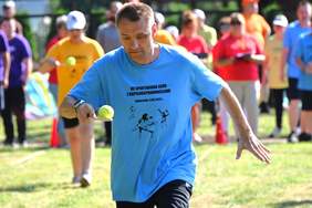 Pokaż zdjęcie: Mężczyzna w jednokolorowej koszulce z napisem VII spartakiada osób z niepełnosprawnościami trzyma w ręku łyżkę na której znajduje się piłka tenisowa.