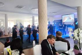 Pokaż zdjęcie: Sala bankietowa z kolumnami. Na środku sali grupa osób trzymająca się za ręce tańczy w kółku. W tle uroczyście zastawione stoły, przy których siedzi kilka osób.