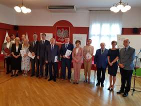 Pokaż zdjęcie: Wspólne zdjęcie uczestników, kilkanaście osób stojących obok siebie w sali konferencyjnej na tle godła, niektóre osoby trzymają w rekach teczki z podpisanymi dokumentami